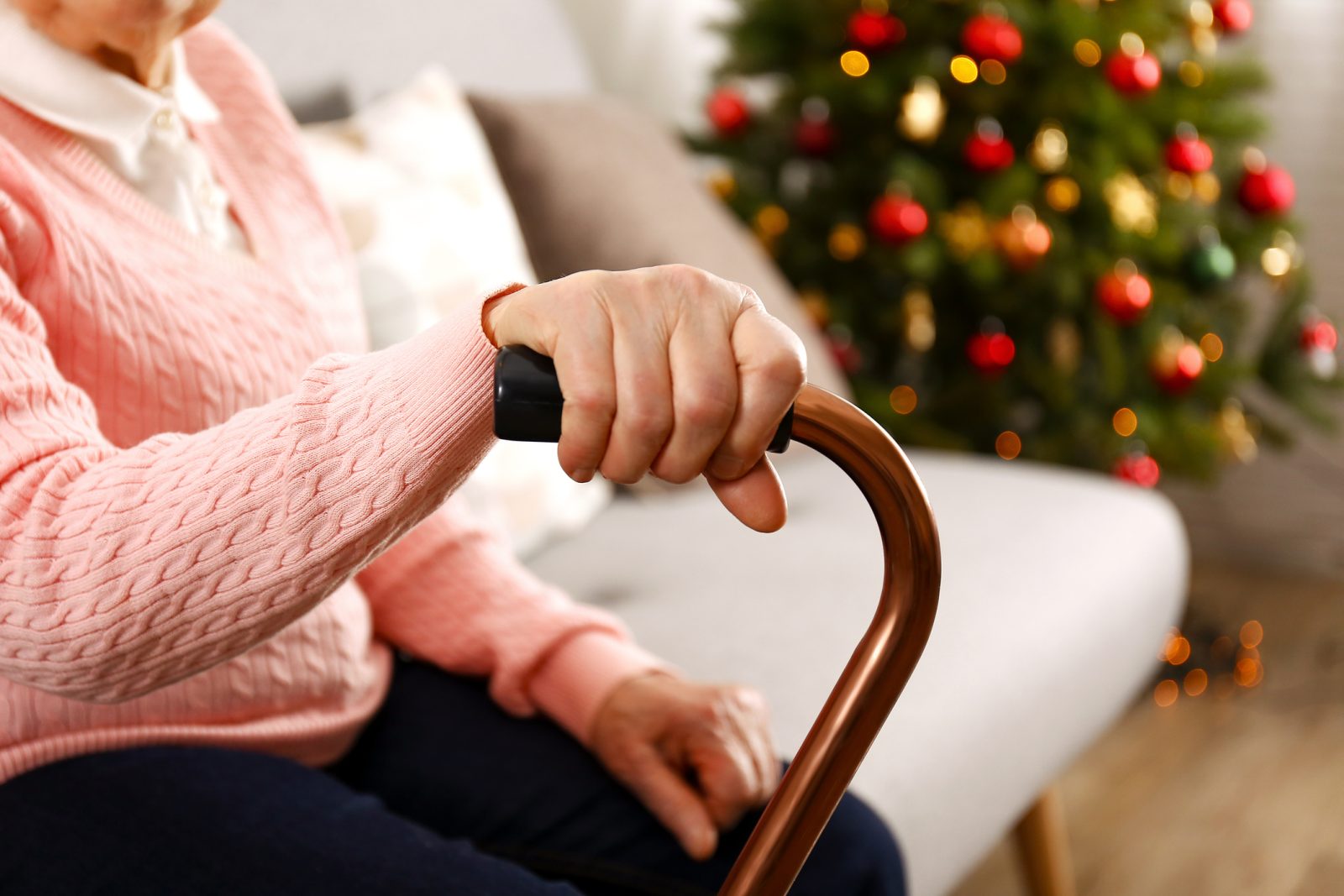 Closeup of senior woman’s hand on a cane while she sits in her living room in front of a Christmas tree.