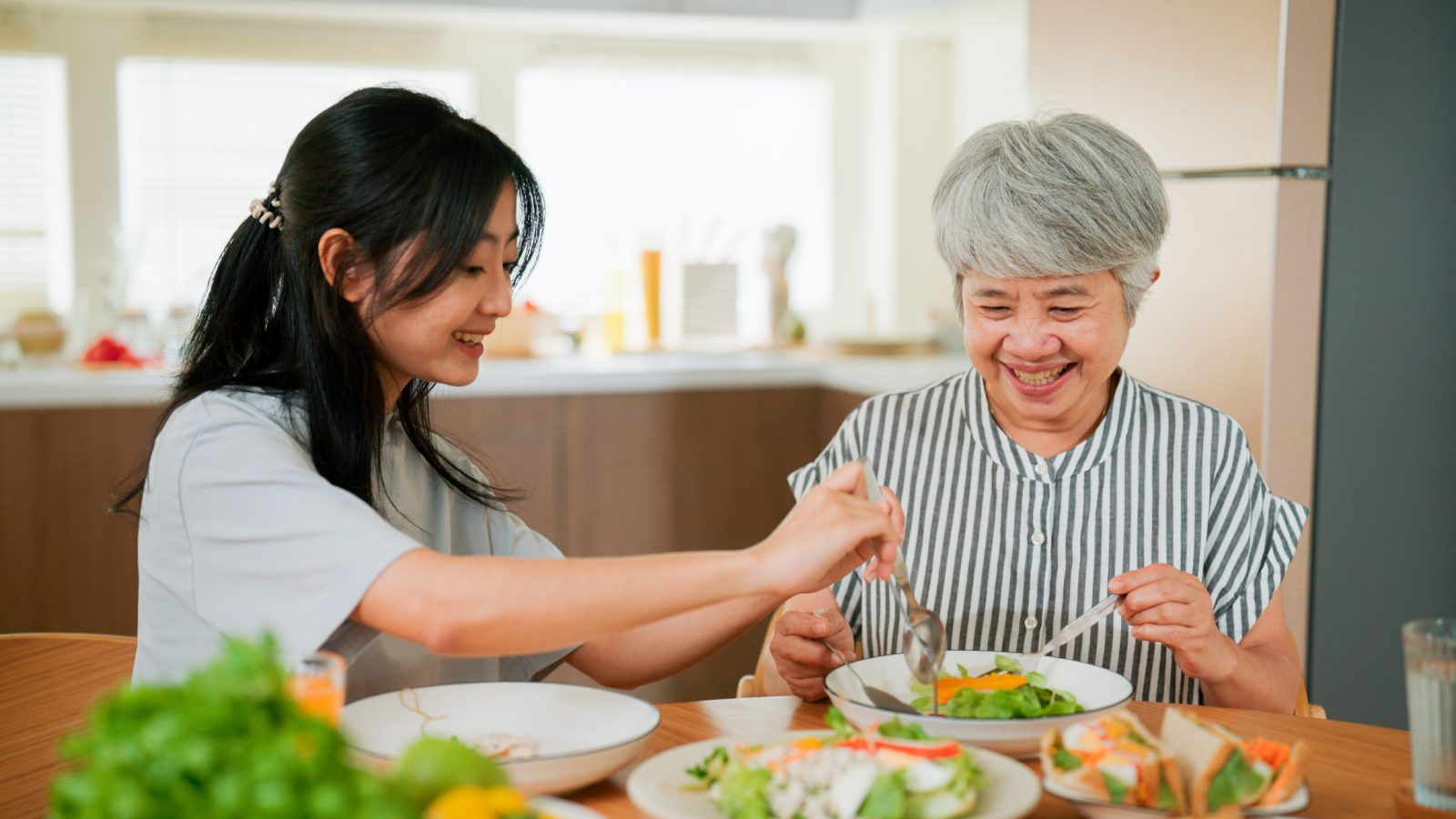 A woman serves healthy food to her senior mother before eating together in their kitchen.