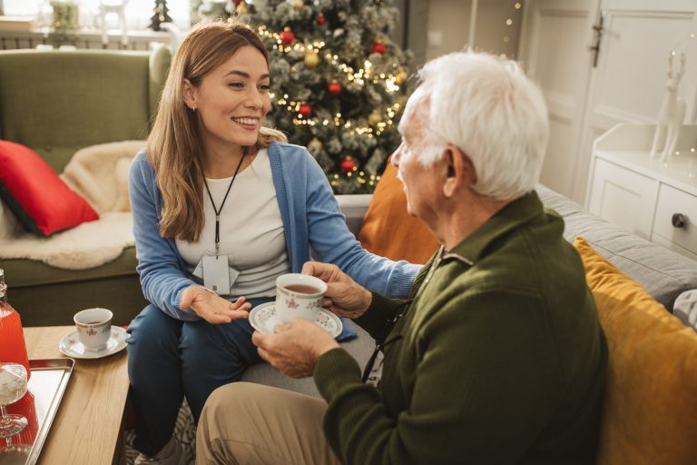 Adult woman who is an in-home caregiver sits with a senior man in his living room. The two share tea in front of his Christmas tree.