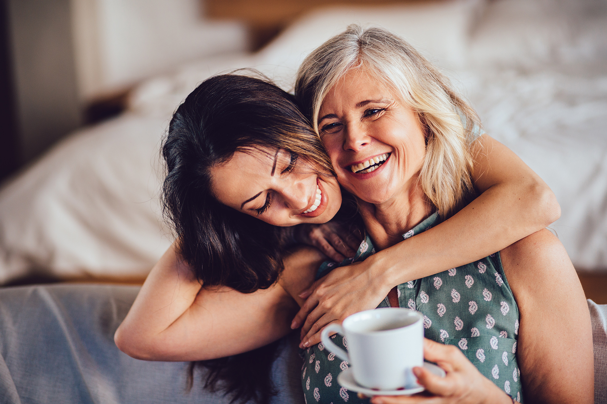 Beautiful adult granddaughter hugging beautiful senior grandmother while sitting on the sofa dinking coffee