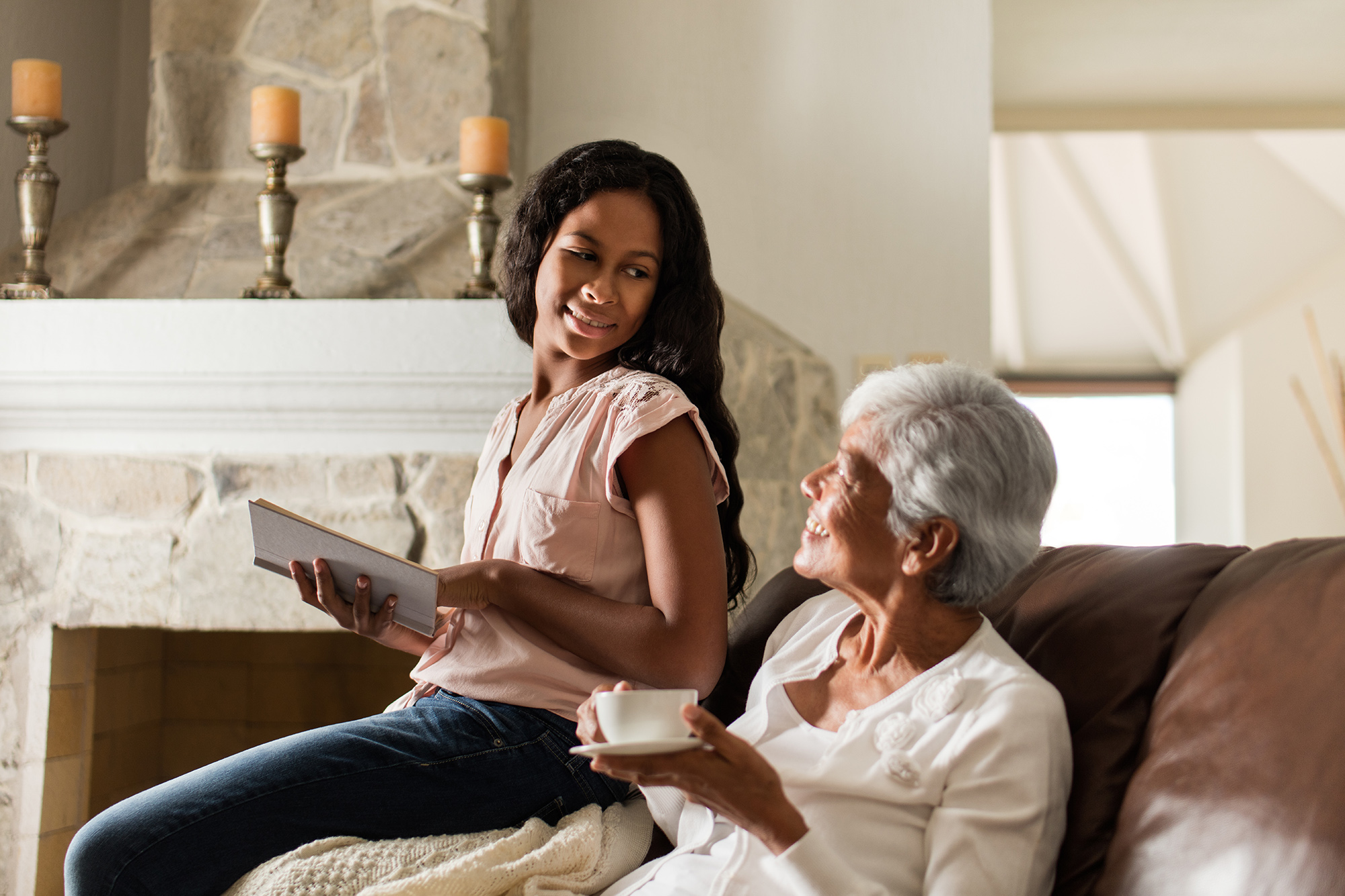 A latin senior woman drinking a cup of a hot beverage on the sofa, sitting next to her granddaughter who is reading a book and smiling at each other.