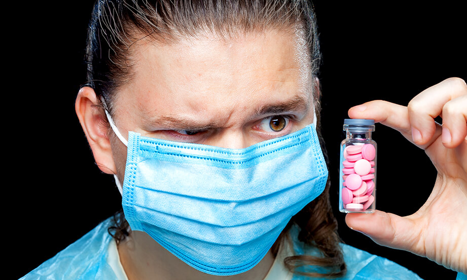 man-in-mask-one-eye-raised-looking-at-a-glass-medicine-jar-with-pink-pills-in-it-sm