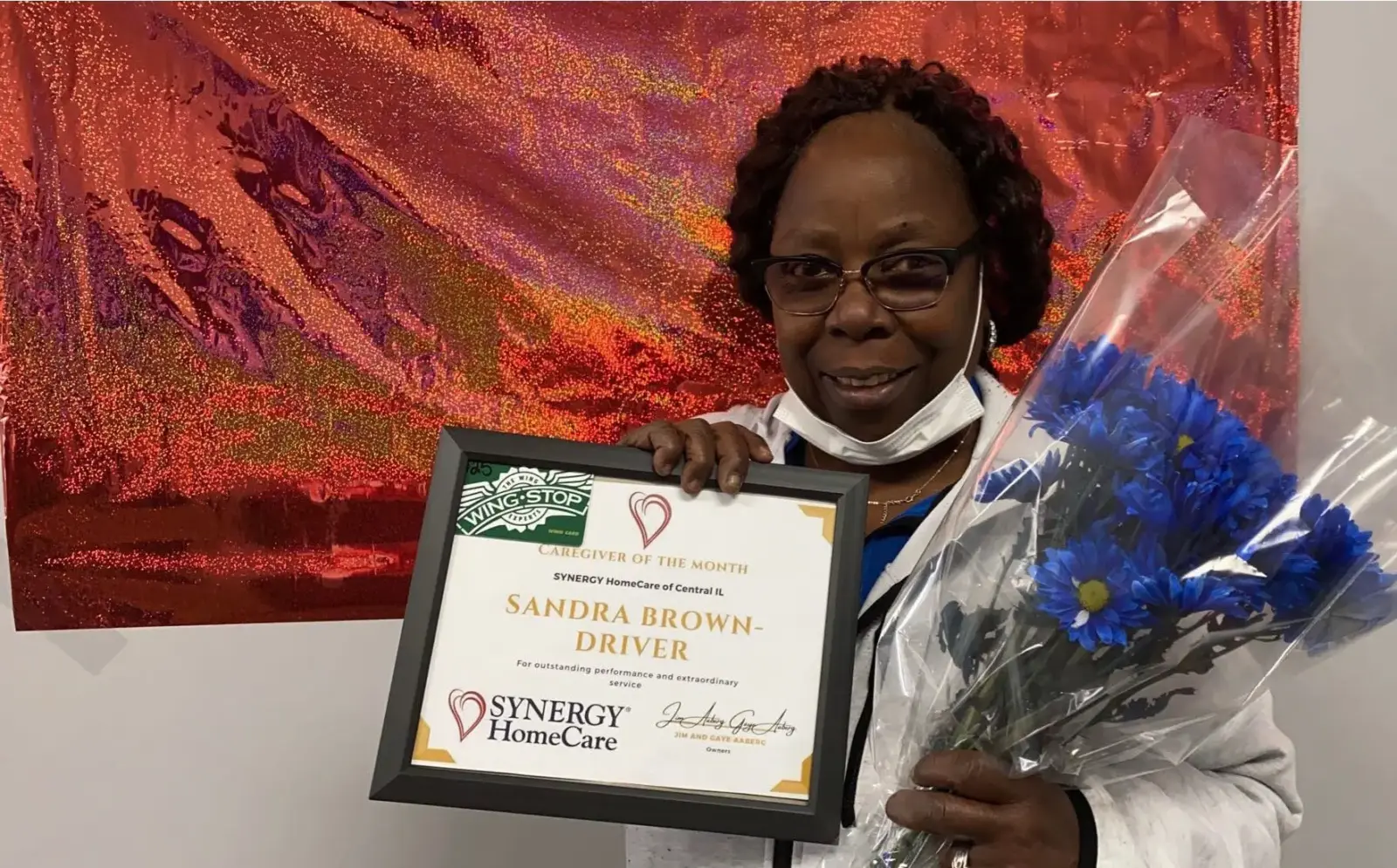 Caregiver of the month Sandra Brown smiles beautifully while hold her awrd certificate and a bunch of beautiful flowers