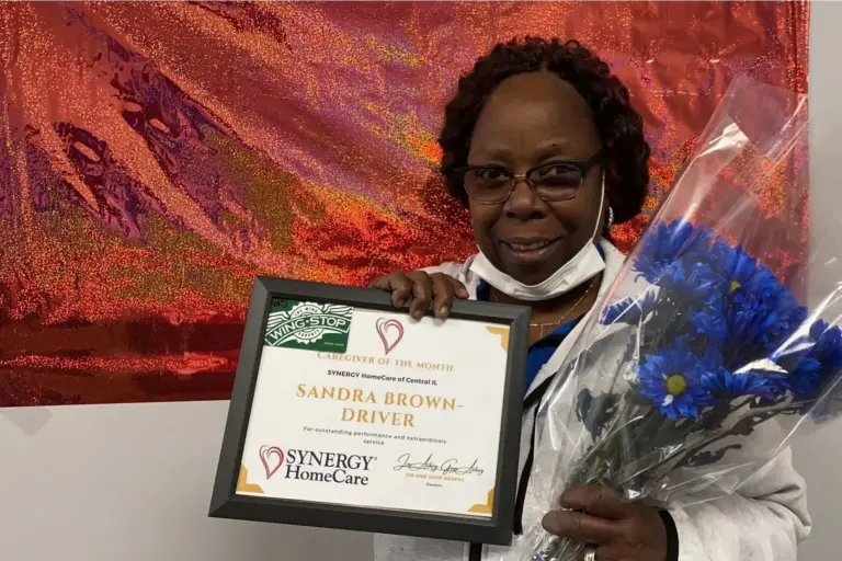 Caregiver of the month Sandra Brown smiles beautifully while hold her awrd certificate and a bunch of beautiful flowers