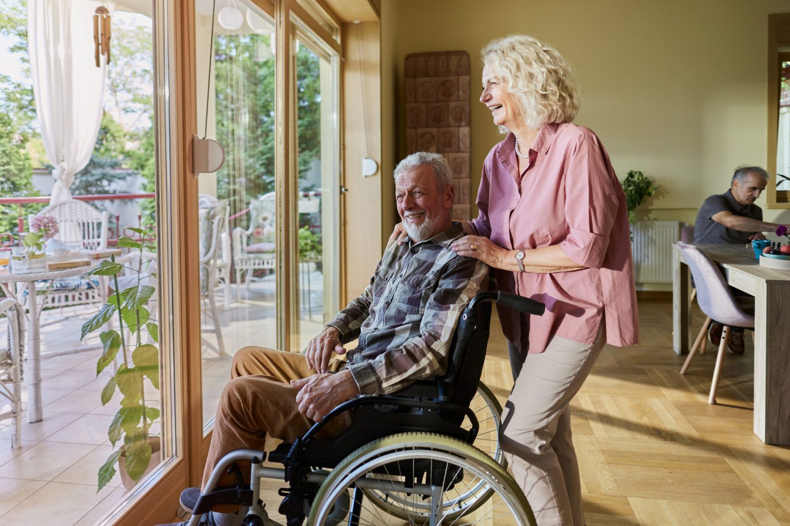Senior man with Parkinson’s disease sits in wheelchair, smiling and looking out the window with his wife who is standing behind his wheelchair and also smiling, looking outside the window.