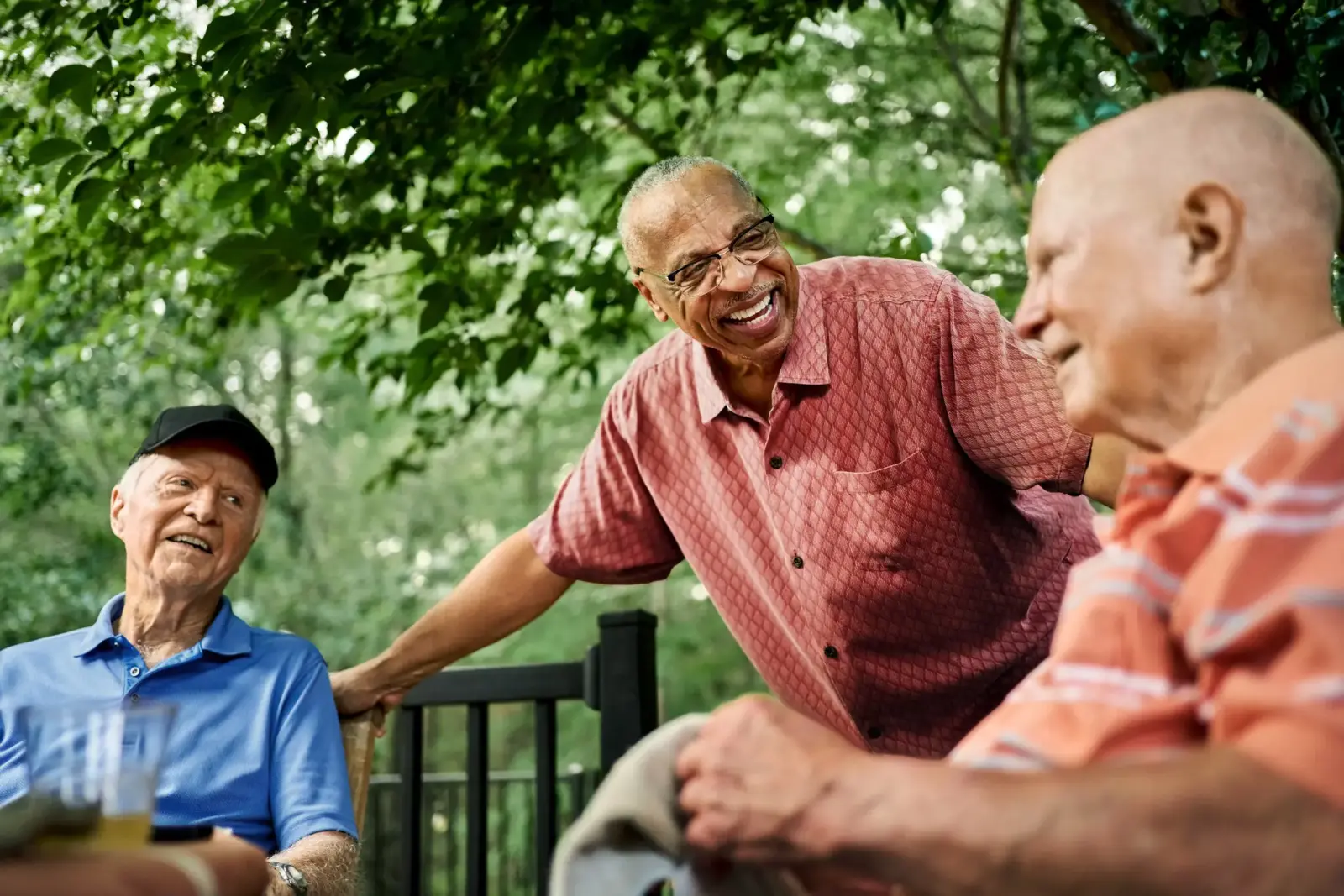 Three happy senior men gathered around a table in an outdoor community space, engaging in conversation and enjoying the serene nature.