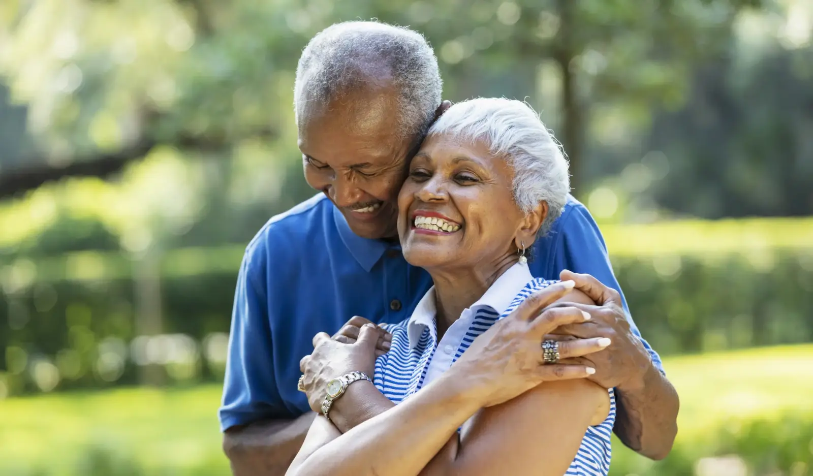 A Santa Monica senior African-American couple, in their 70s, standing together at the park, surrounded by lush foliage. The man is hugging his wife from behind and she is grinning.