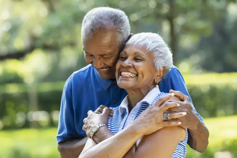 A Santa Monica senior African-American couple, in their 70s, standing together at the park, surrounded by lush foliage. The man is hugging his wife from behind and she is grinning.