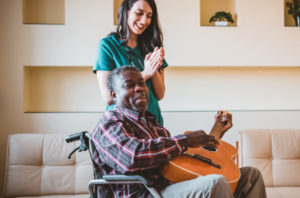 senior man in wheelchair playing the guitar while SYNERGY caretaker claps 