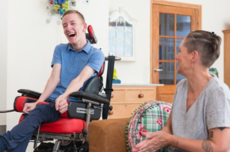 A disabled young adult male sitting in his wheelchair and laughing with a friend.